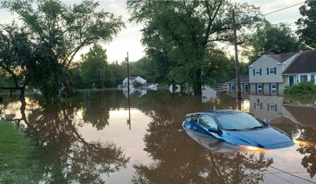A heavily flooded road in a Hillsborough neighborhood, with water submerging the lower half of a car, leaving only the roof and upper windows visible.