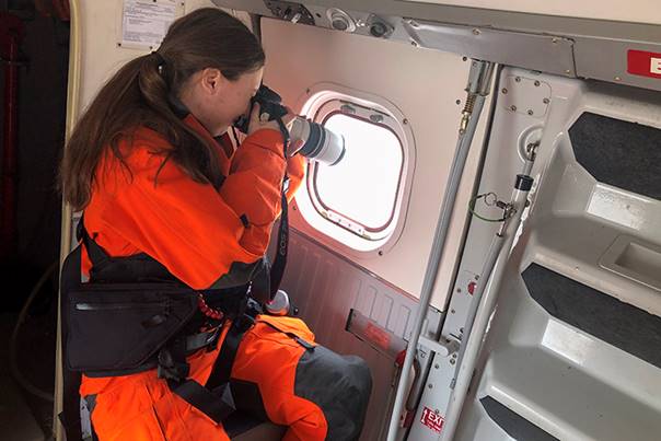 A woman wearing an orange flight suit holds a camera up to a plane’s window to take a photo.
